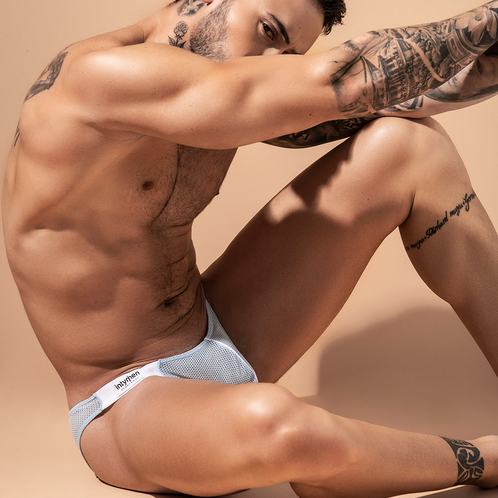 A tattooed person sits on a light surface wearing Intymen Fancy Brief. The low-waist mens underwear highlights their form as they rest an arm on their knee, gazing downward against a neutral beige background.