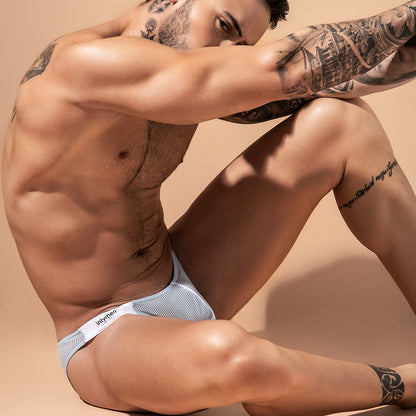 A tattooed person sits on a light surface wearing Intymen Fancy Brief. The low-waist mens underwear highlights their form as they rest an arm on their knee, gazing downward against a neutral beige background.