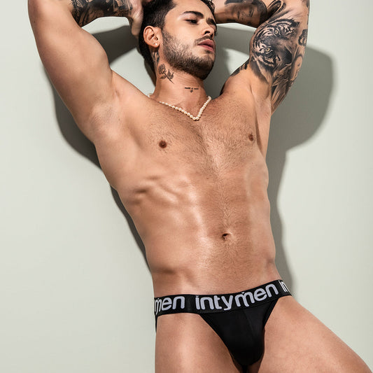 A man poses with tattooed arms raised, wearing Intymen Peace Brief made from stretchable cotton for comfort, featuring a supportive pouch design. Against a neutral backdrop, his necklace stands out as he showcases the black underwear.