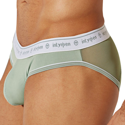 A person models the Intymen See Through Back Brief in light green, styled with breathable cotton and featuring white mesh accents on the sides. The waistband showcases Intymen repeatedly in white. The image captures a side view with subtle transparency details.