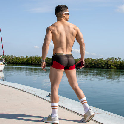 A man exudes sporty appeal on a waterfront walkway wearing Pistol Pete Athletic Wide Waistband Boxer Trunks in red and black, white athletic socks, sneakers, and sunglasses. The clear sky frames him as a boat glides by in the distance.