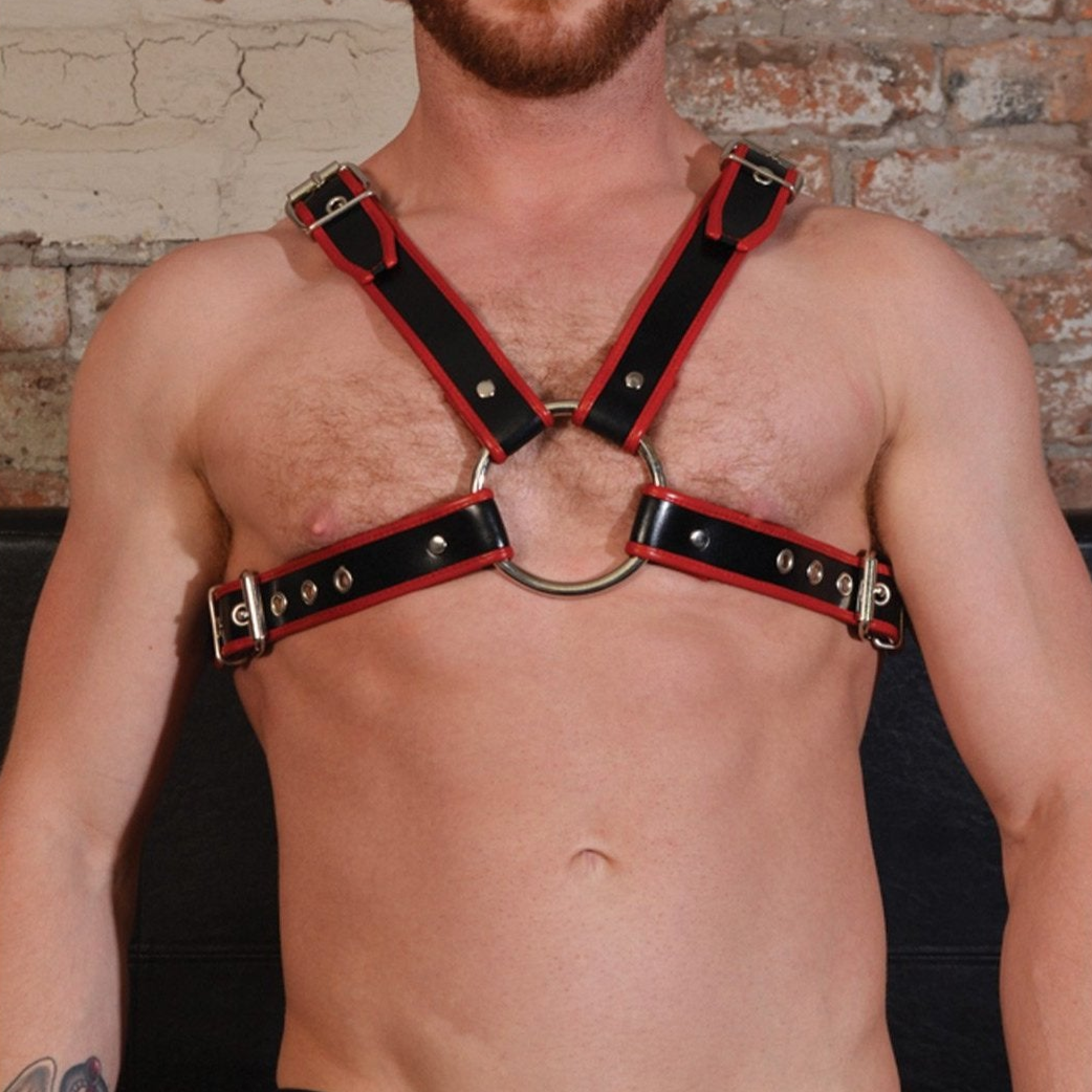 A shirtless person models the Rouge Chest Harness from Adams Toy Box—black and red with buckles and a circular chest ring. Tattoos on their arm and a beard add flair as they stand against a brick wall.