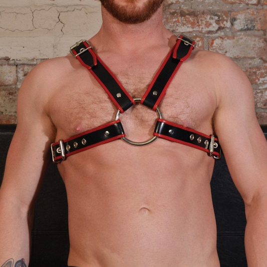 A shirtless person models the Rouge Chest Harness from Adams Toy Box—black and red with buckles and a circular chest ring. Tattoos on their arm and a beard add flair as they stand against a brick wall.