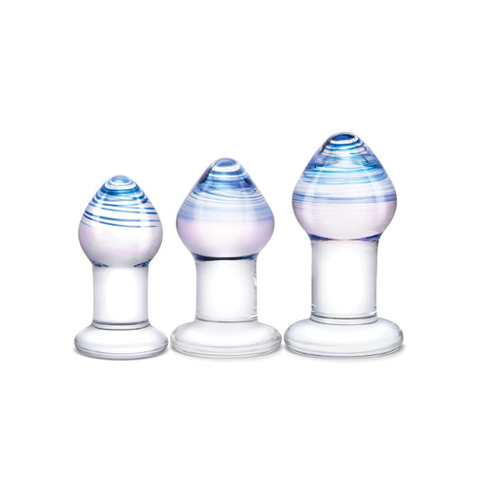 The Gläs Pleasure Droplets Anal Training Kit includes three glass butt plugs of different sizes, featuring a clear base and rounded top with light blue and pink swirls. The transparent glass softly reflects light, creating an enchanting look.