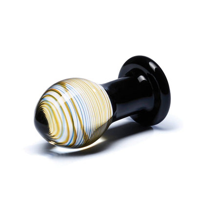The Gläs Galileo Glass Butt Plug, made from pyrex glass in an elongated orb shape, features swirling white, blue, and yellow patterns with a black handle-like base. This striking piece rests boldly on a plain white surface.