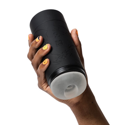 A hand holds the ROMP Dash Stroker, a black cylinder with textured patterns and a translucent accent, made from body-safe silicone. The nails, painted with smiley faces, complement this skin-soft product against a white backdrop.