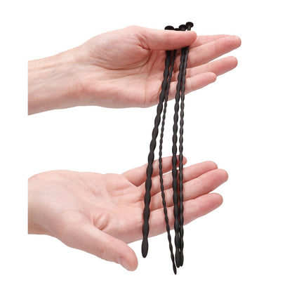 Two hands hold long black twisty silicone plugs from the Shots Ouch Beginner Urethral Sounding Silicone Spiral Screw Plug Set against a plain white background, evoking a beginner kit layout.