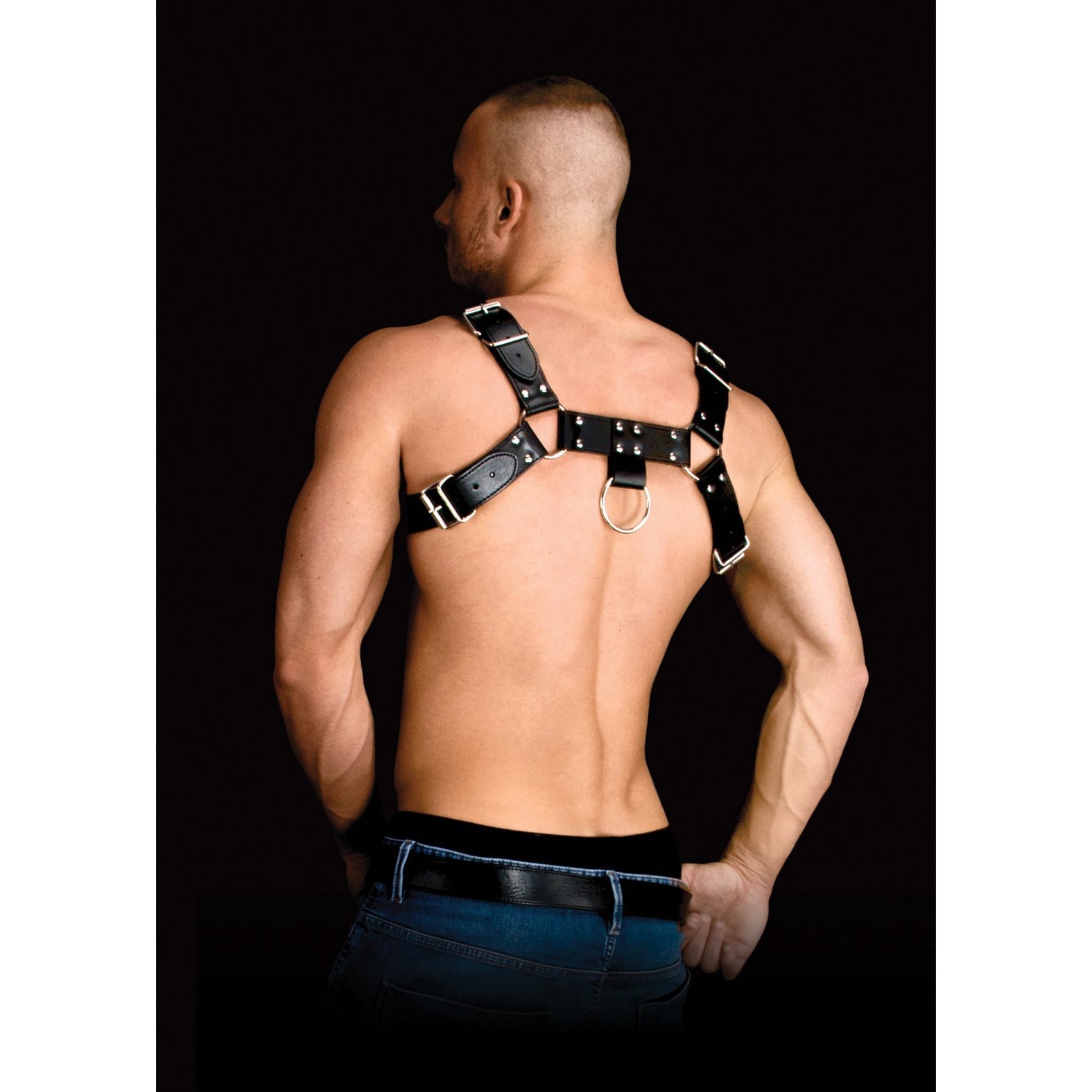 A man confidently showcases the Shots Ouch Costas Solid Structure 2 Body Harness. Hes shirtless, with short hair and hands on his hips, set against a black background that accentuates its sleek design and strong material.