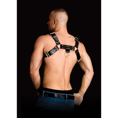 A man confidently showcases the Shots Ouch Costas Solid Structure 2 Body Harness. Hes shirtless, with short hair and hands on his hips, set against a black background that accentuates its sleek design and strong material.