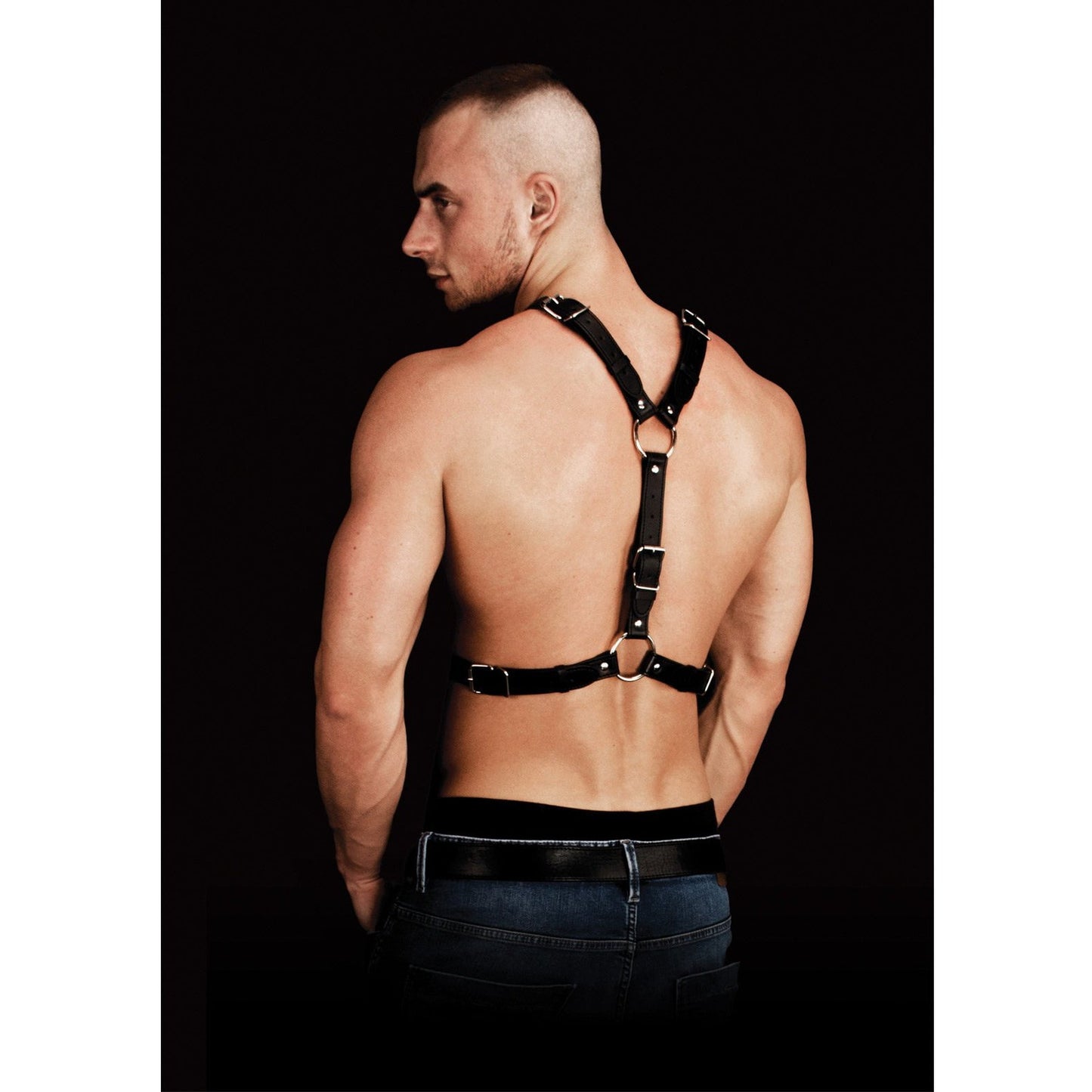 A muscular man with a shaved head models the Shots Ouch Thanos Chest Centerpiece Body Harness, its bonded leather design enhancing his physique. Shirtless and in dark jeans, he faces away, the shadowy backdrop emphasizing both the craftsmanship of the harness and his toned body.