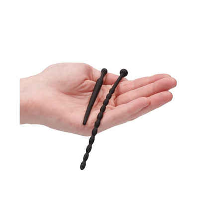 A hand displays two beginner-friendly black silicone urethral sound plugs from the Shots Ouch Urethral Sounding Beginners Set by Adams Toy Box. One is smooth and tapered for gentle exploration, while the other features a beaded texture for variety, set against a white background.