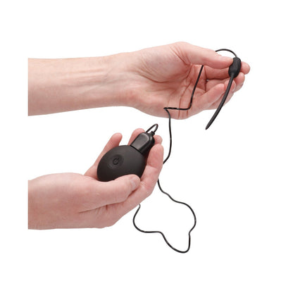 Hands holding a small black Shots Ouch Vibrating Plug with a wire and ear hook. The wire connects to a round, black device with a button, resembling the sleek design of this silicone rechargeable gadget. Background is plain white.