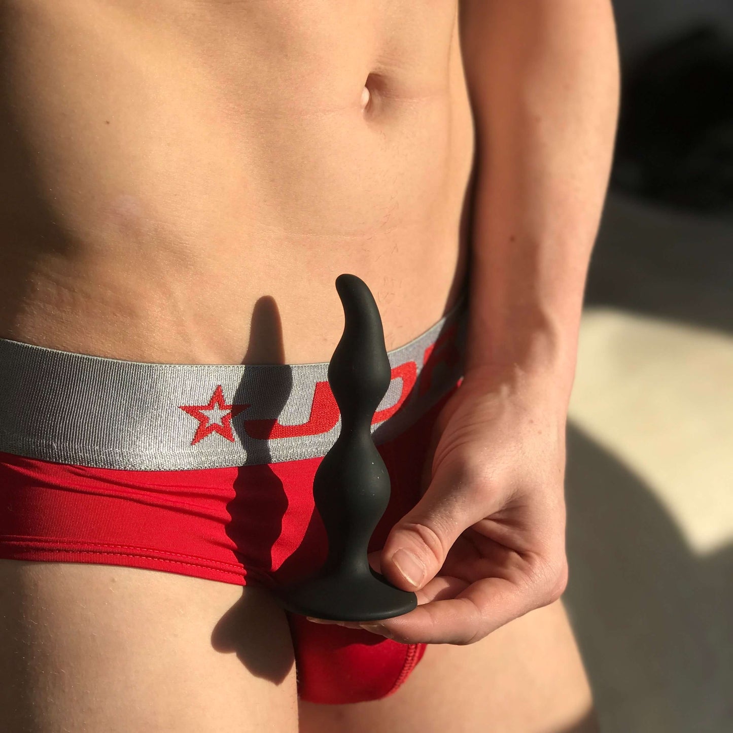 A person wearing red underwear holds a wavy-shaped silicone toy from the Zero Tolerance Intro To Prostate Kit. The softly blurred background accentuates both the object and clothing, suggesting a moment of P-spot pleasure.