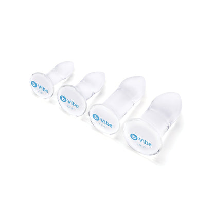 The b-Vibe Glass Anal Dilator 4 pc Set features four transparent, graduated silicone plugs with round bases labeled b-Vibe, arranged by size on a white background and designed for temperature play. Made from hypoallergenic, body-safe materials, they offer safe and pleasurable versatile sizing.