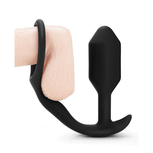 The b-Vibe Snug & Tug Weighted Silicone & Penis Ring, a black silicone toy with an insertable piece and loop to enhance prostate orgasms, is displayed on a light-colored abstract surface.