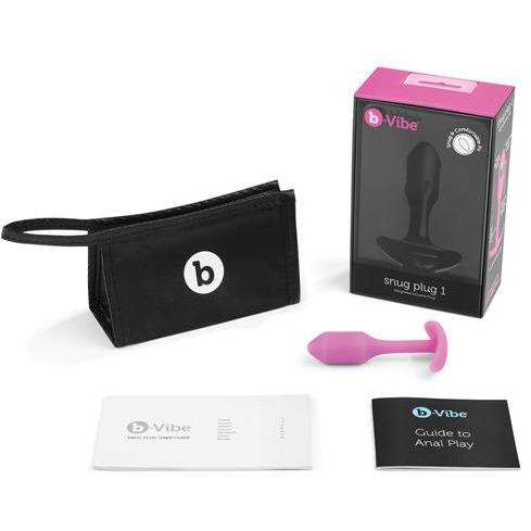 The B-Vibe Snug Butt Plug 1 is elegantly displayed on a white surface, featuring a pink 100% silicone plug with a weighted base, alongside a black storage pouch, box with product image and logo, and two booklets: one with the b-Vibe logo and another titled Guide to Anal Play.