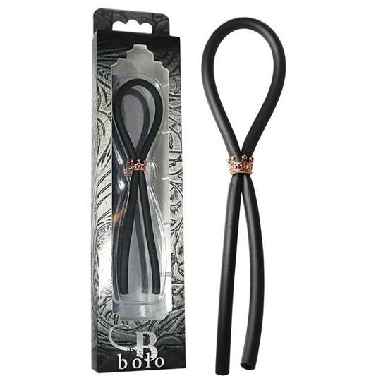 Adams Toy Box presents the Bolo Silicone Lasso with a rose gold crown slider ring, neatly displayed in decorative packaging with black and gray abstract designs. A clear window reveals the elegant black silicone tie crafted from body-safe materials.