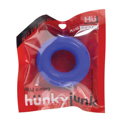 The image showcases a blue Hunky Junk C Ring from the Singles collection in a red and transparent plastic package, labeled hunkyjunk and HUJ c-ring by Hunky Junk. This silicone cock ring features playful packaging with creative branding text.