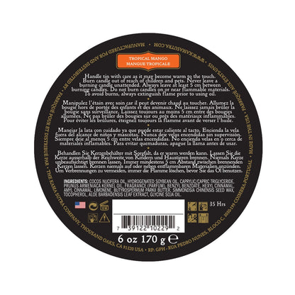 A black circular lid on the Kama Sutra Ignite Massage Candle in Coconut Pineapple shows multilingual safety info, product details, weight (6 oz/170g), 35-hour burn time, and moisturizing formula. It also displays a barcode, manufacturer details, and scent of tropical mango.