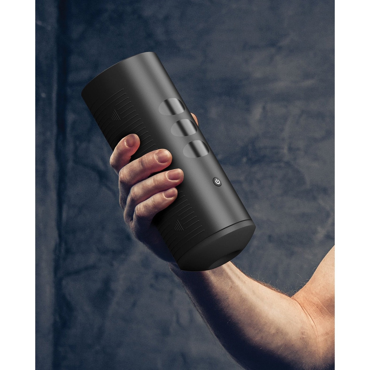 A person holds a Kiiroo Titan Interactive Vibrating Stroker, a black cylindrical device with an ergonomic design and buttons, against a textured dark surface.