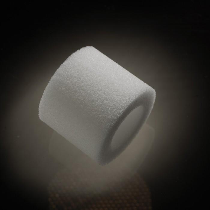 A cylindrical, white, sponge-like object with a hollow center reminiscent of the Male Edge Pro Penis Enlarger Kit by Male Edge is set against a dark background. Soft illumination from above casts a faint shadow beneath.