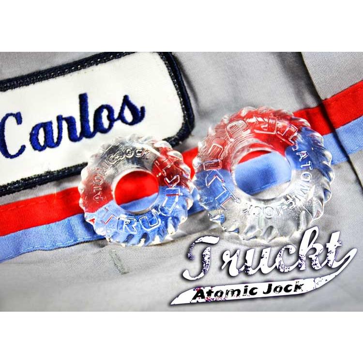 Close-up of two clear Skinflex TPR rings on a gray fabric with red and blue stripes, featuring a label that reads Carlos and brand identifiers Truckt and Atomic Jock, emphasizing the Oxballs TruckT Cock & Ball Ring - Pack of 2.