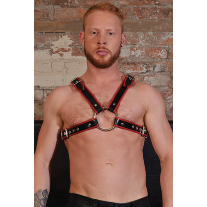 A man with short hair and a beard confidently dons the Rouge Chest Harness by Adams Toy Box, a chic BDSM accessory, against a brick wall backdrop.