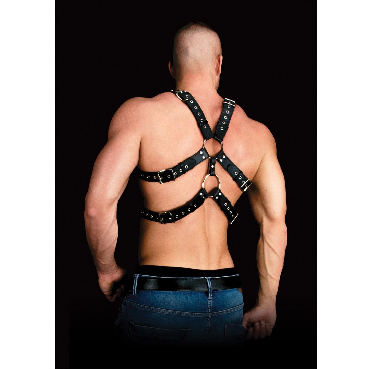 A man models the Shots Ouch Andreas Masculine Masterpiece Body Harness by Adams Toy Box. The adjustable black leather harness features metal rings and buckles, paired effortlessly with jeans against a plain black backdrop, highlighting its bonded leather design.